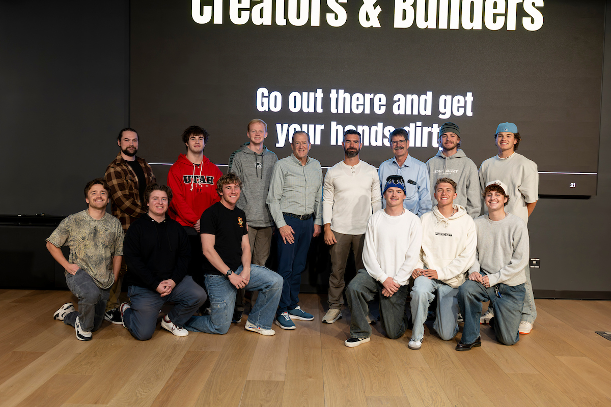 A group photo on stage featuring speaker, McKay Christensen and attendees. Behind them, a large screen displays the text: “The Country Needs Creators & Builders. Go out there and get your hands dirty.”