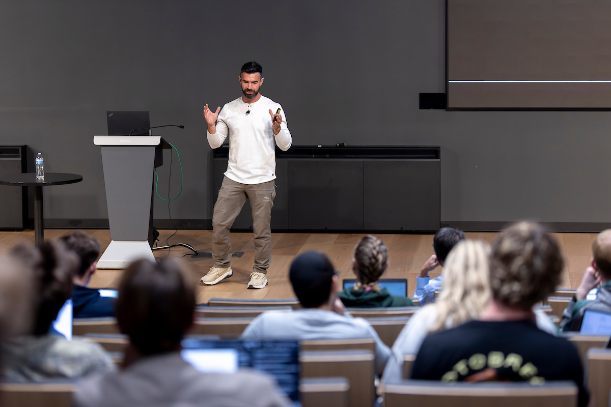 McKay Christensen presents to an audience, gesturing with both hands. The audience is seated in tiered rows with laptops open, and a podium is visible to the left.