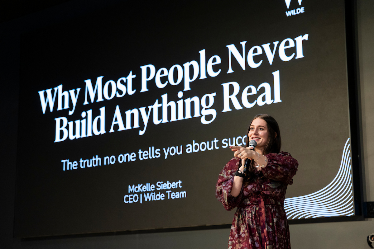 McKelle Siebert speaking on stage in front of a slide titled “Why Most People Never Build Anything Real.”