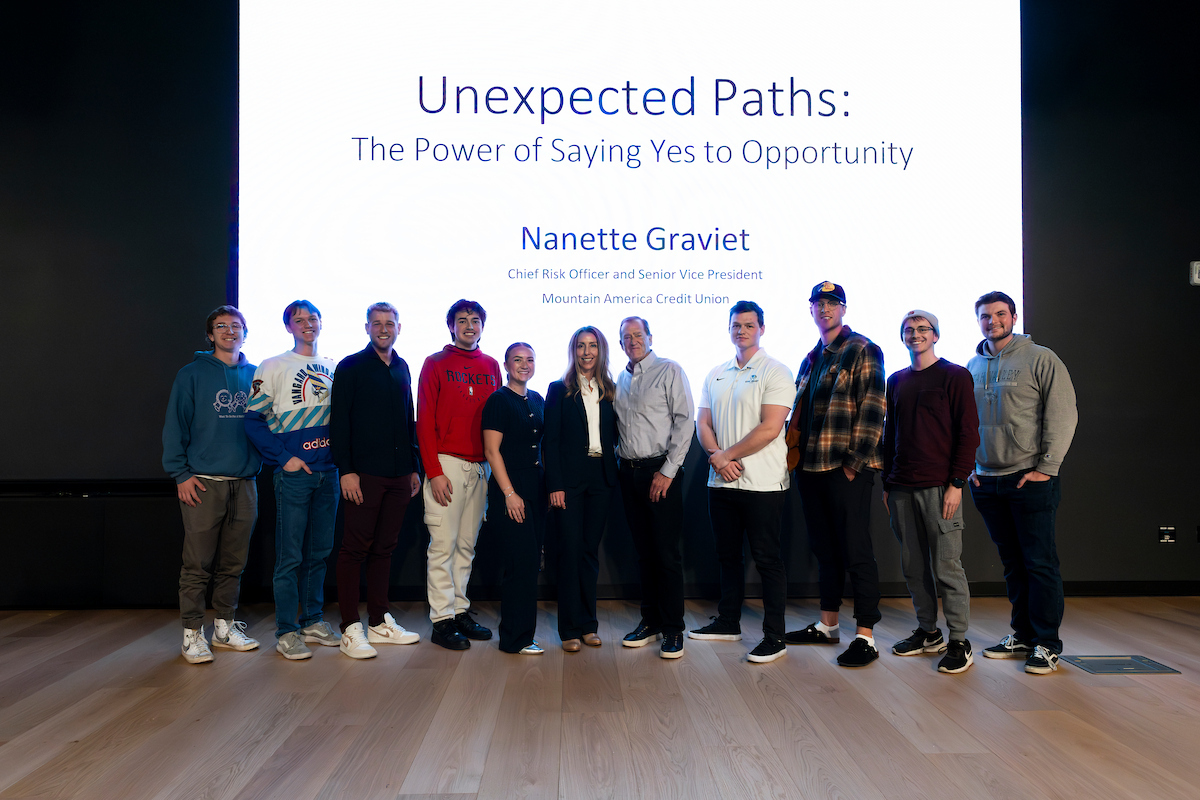 Group photo of Nanette Graviet with UVU students on stage in front of a large presentation screen displaying “’Unexpected Paths: The Power of Saying Yes to Opportunity’ by Nanette Graviet.” 