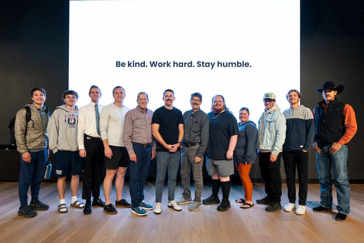 Taylor Napierski stands with UVU students and faculty in front of the Halladay Lecture Series screen displaying his motto: “Be kind. Work hard. Stay humble.”