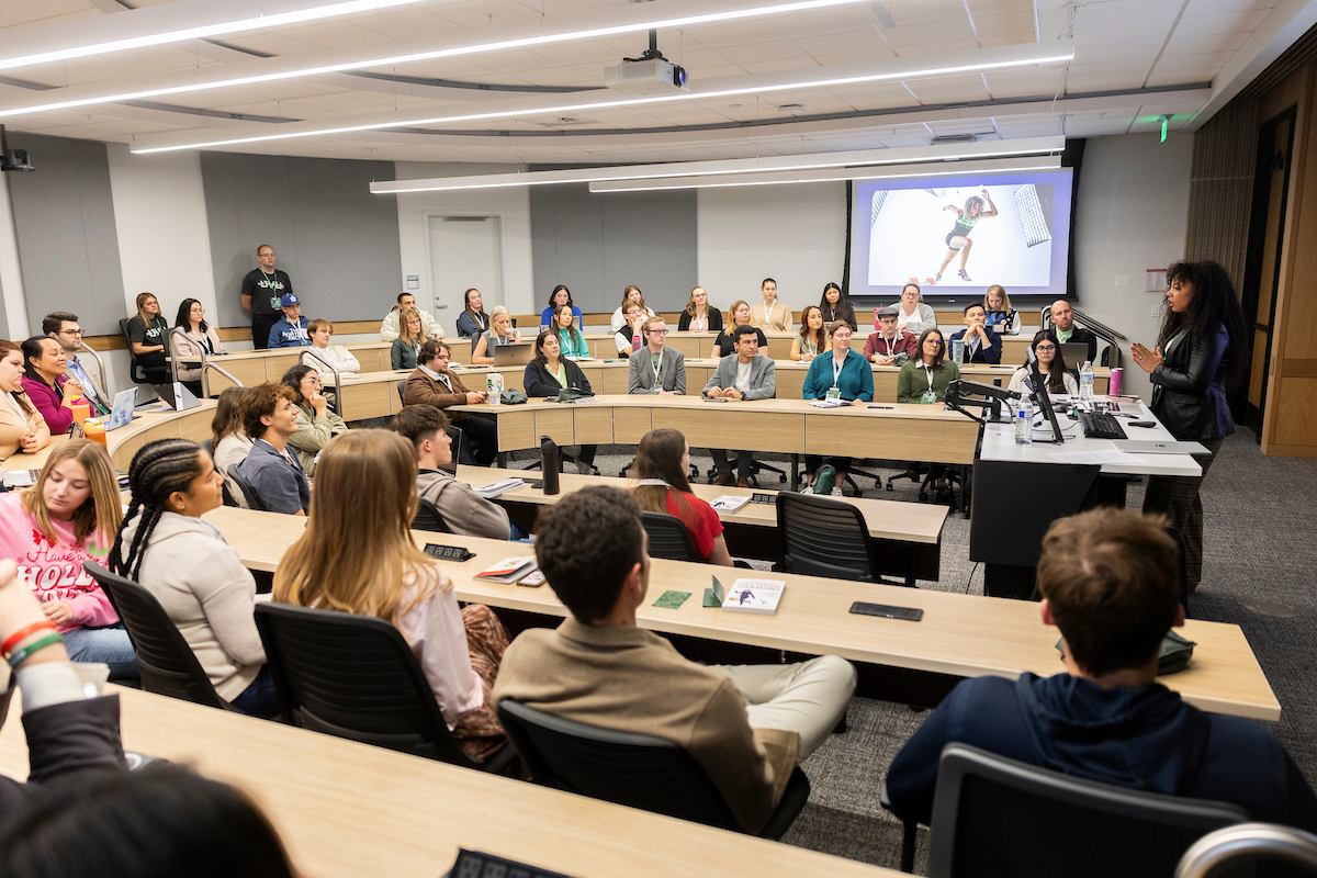 Students and professionals seated in a tiered classroom listening to a speaker presenting at the front of the room.