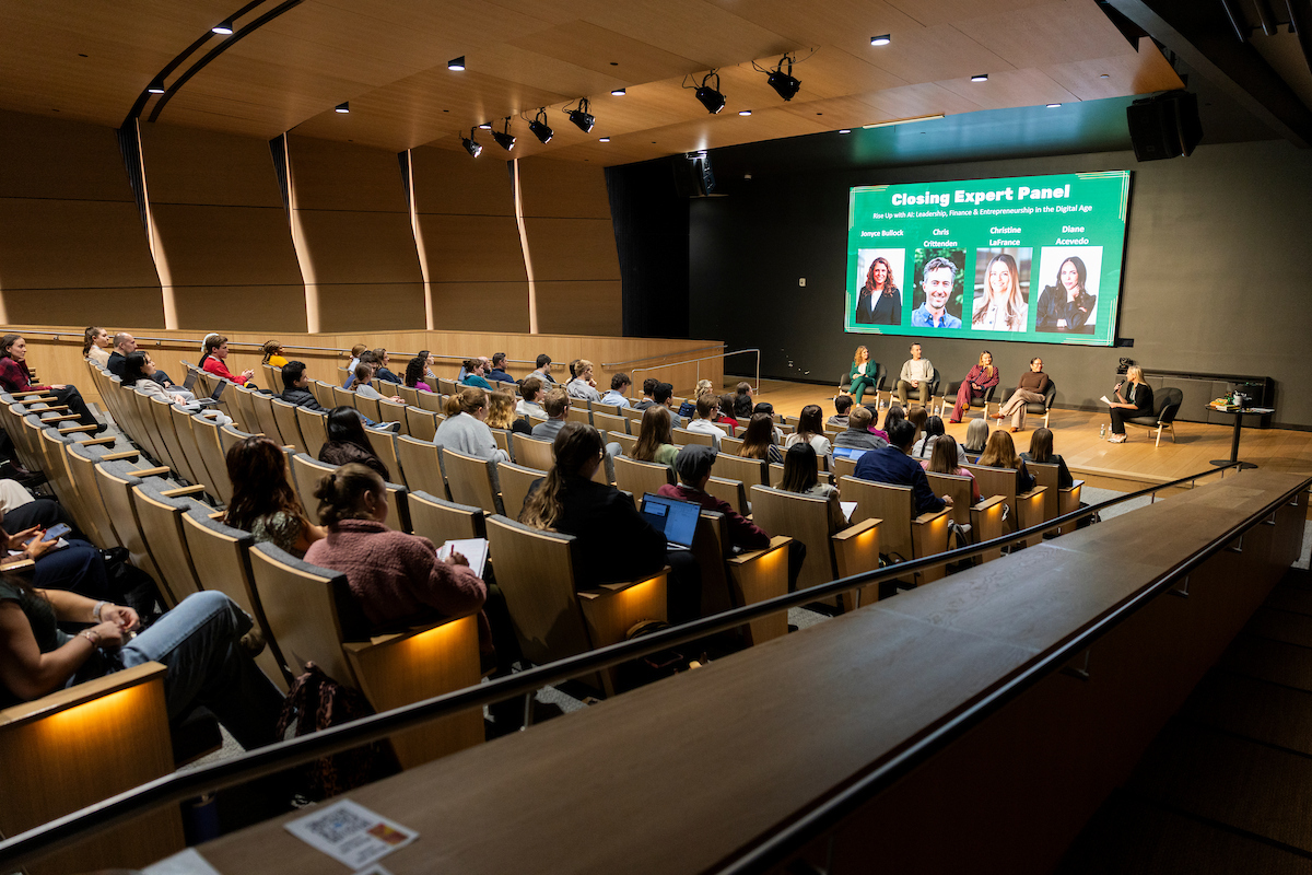 Audience seated in an auditorium watching a panel of experts speak on stage beneath a large screen.