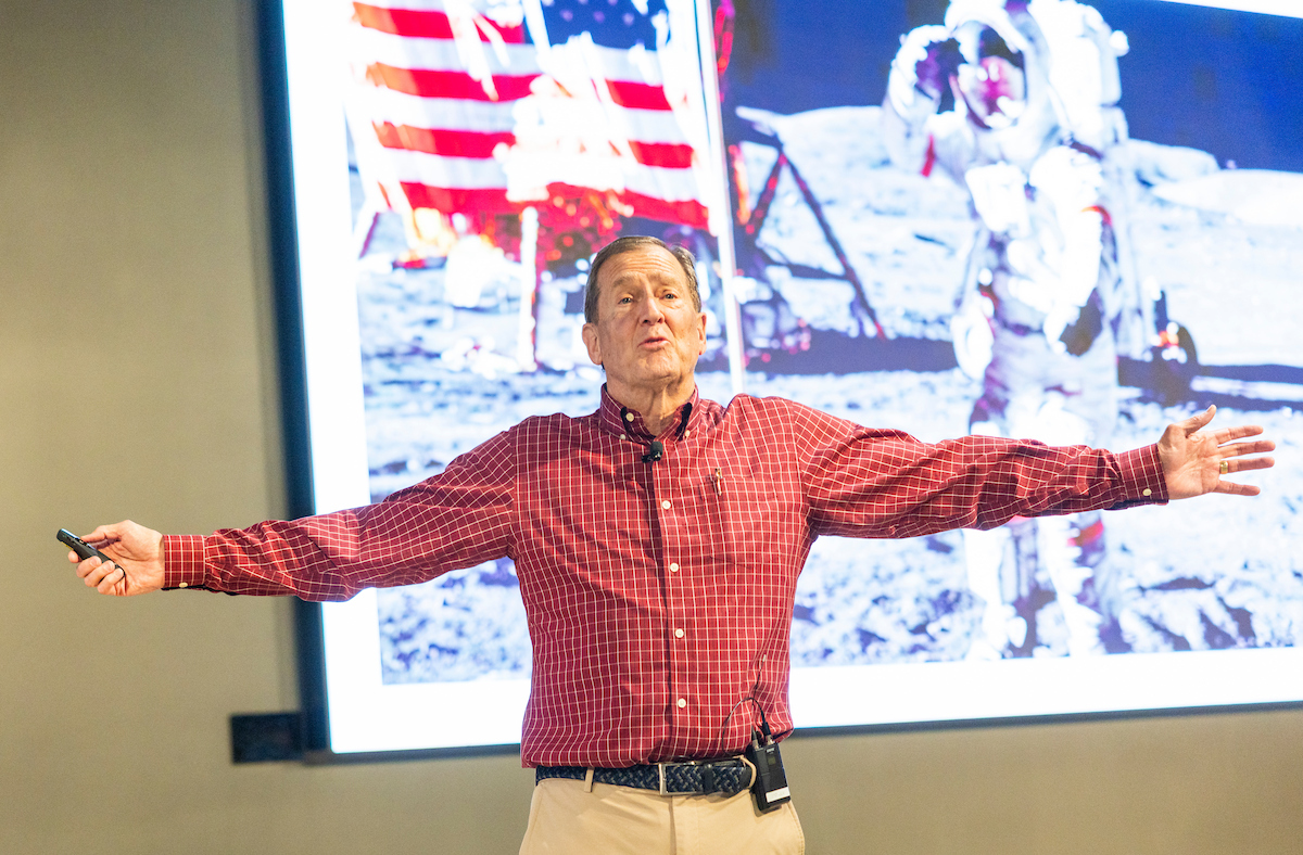 Tom MacDonald speaks passionately at the Fall 2025 Halladay Lecture Series, arms outstretched as he emphasizes a key point with a projected image of the moon landing behind him.