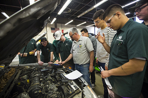 Students standing around an open car hood while a professor teaches them