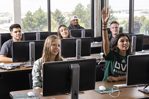 Students behind individual computers sitting in a class as one student raises her hand