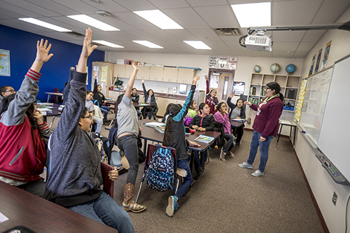 A classroom of students raising their hands and a teacher calling on a student