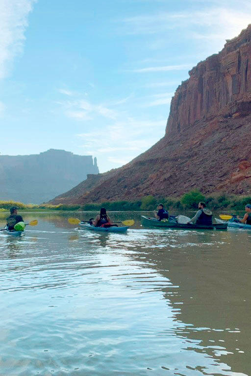 A group of people kayaking away from the camera by red rock cliffs