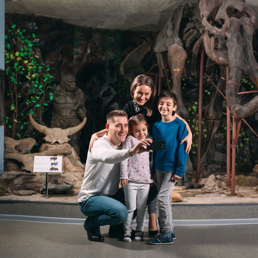 a family of a father, mother, and two children pose for a selfie in front of a dinosaur replica
