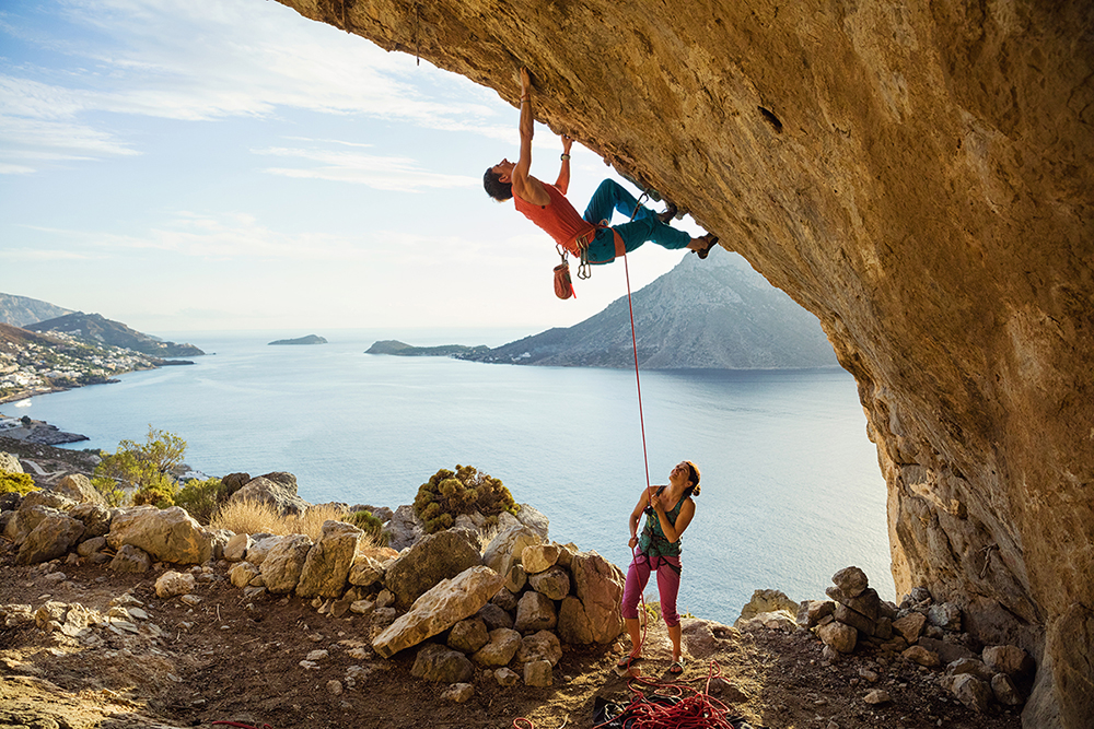 woman holds rope as man climbs rock overlooking a lake