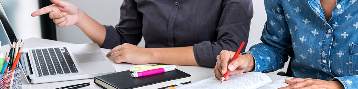 two women looking at laptop with notebooks and paper on desk