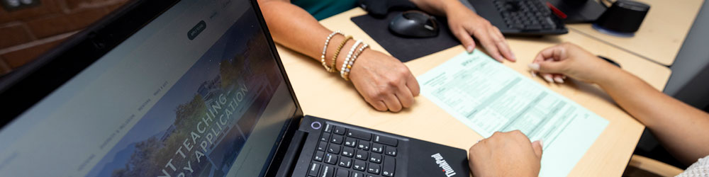 Two people doing paperwork on a desk with a laptop next to them.