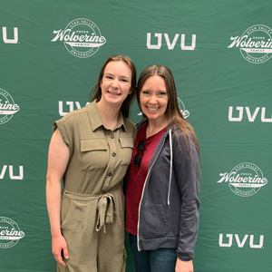 Two women stading next to each other with one arm around each other smiling at the camera standing in front of a UVU backdrop behind them.