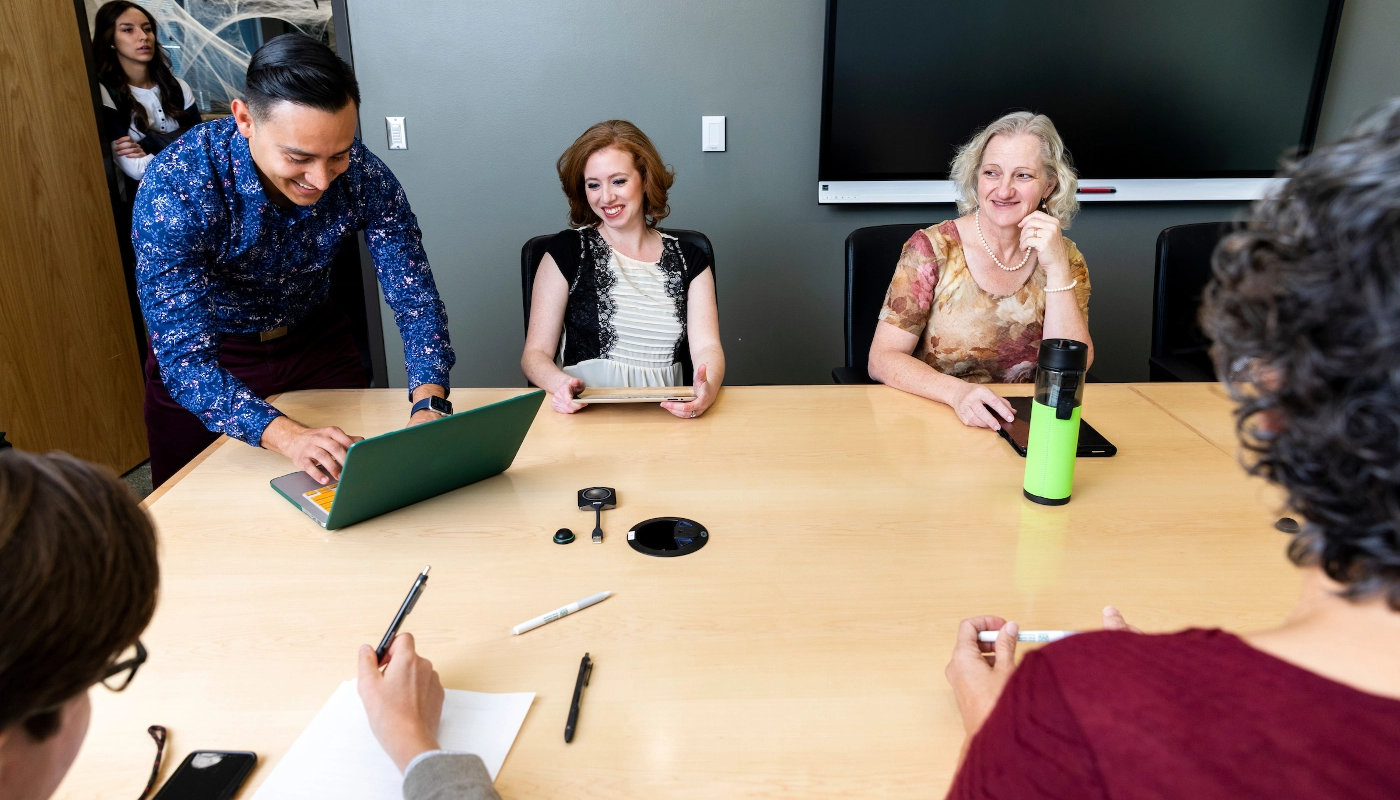 Employees seated around a desk. One employee is working on a laptop.