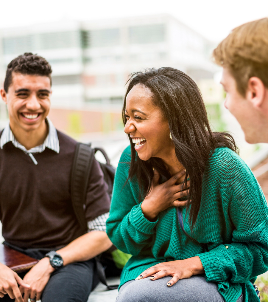 Three students sitting together in conversation. A female student in the middle is laughing.