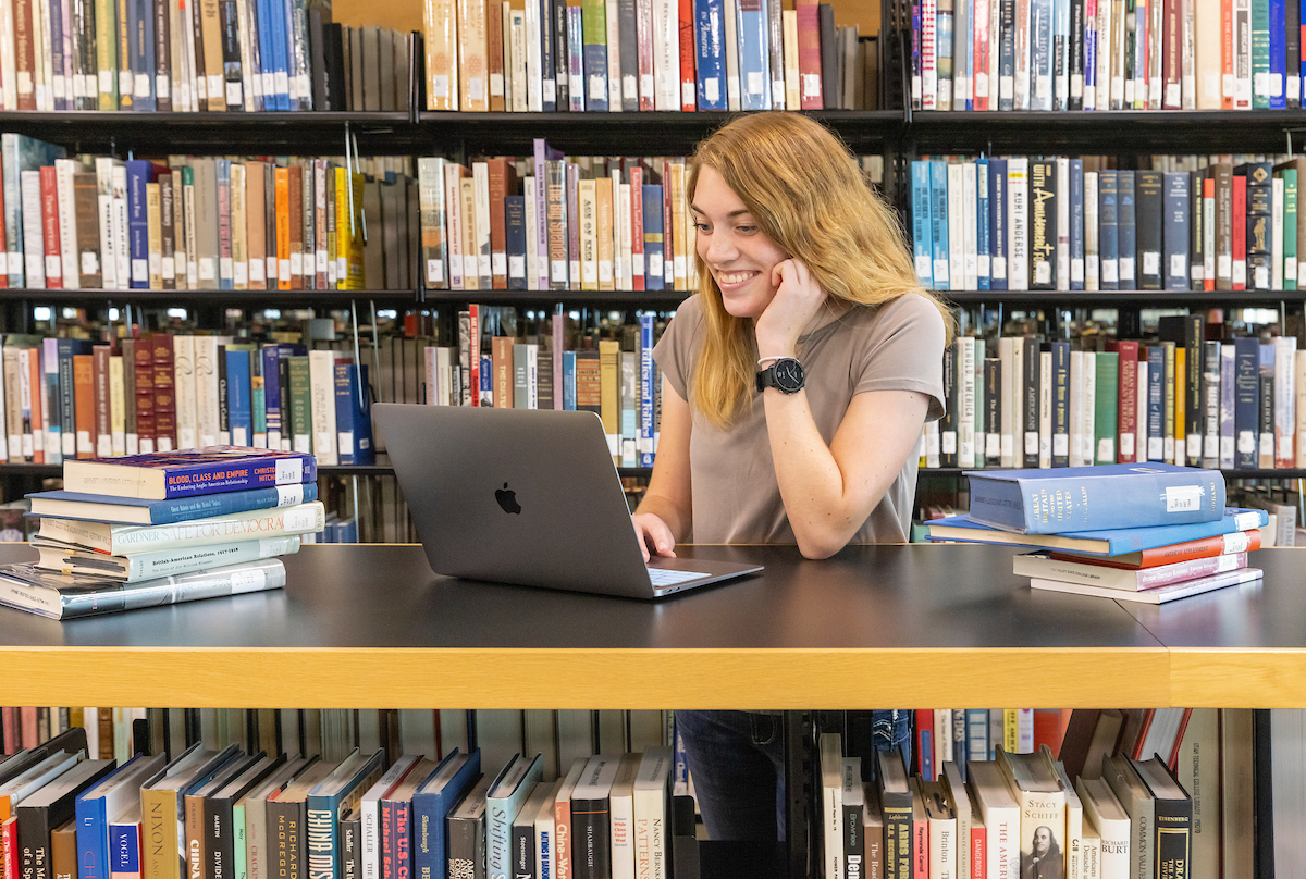 Image of student looking at their laptop, a shelf full of books is behind them
