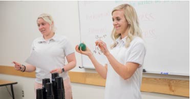 Two female students stand in front of a class showing different massage tools