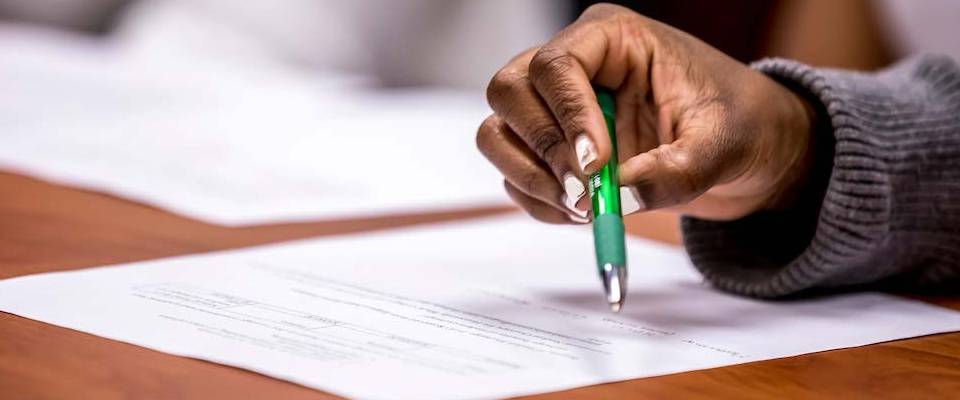 A hand holding a pen about to write on a sheet of paper that is on a desk.