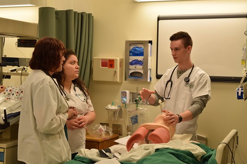A man explains something to two onlookers over a manequin in a hospital bed