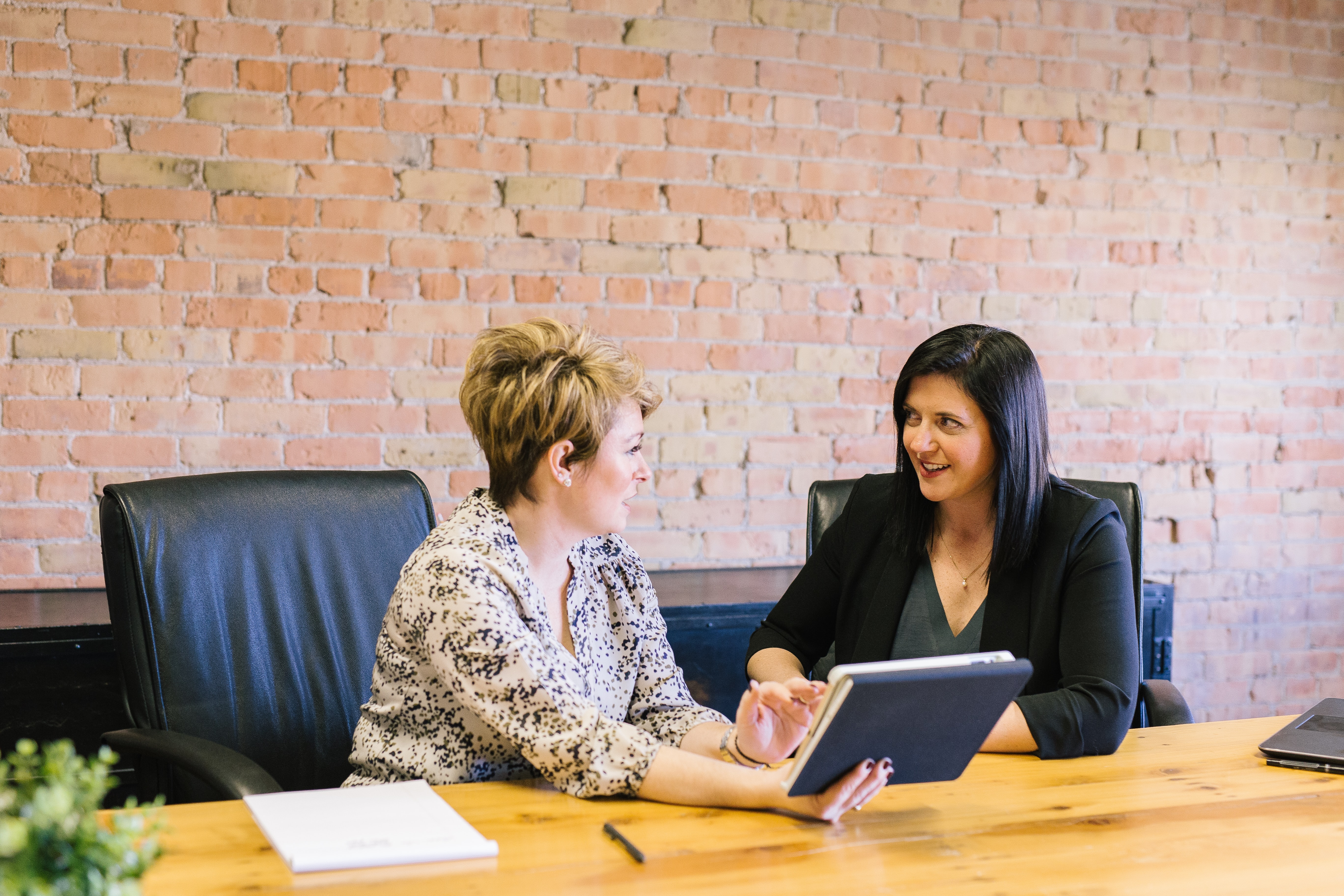 Two women having a conversation at a table. One is taking notes on paper and the other is holding a tablet.