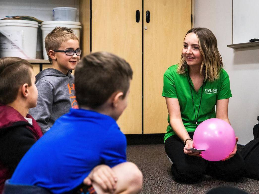 An intern in a green shirt holds a balloon and smiles at children near her