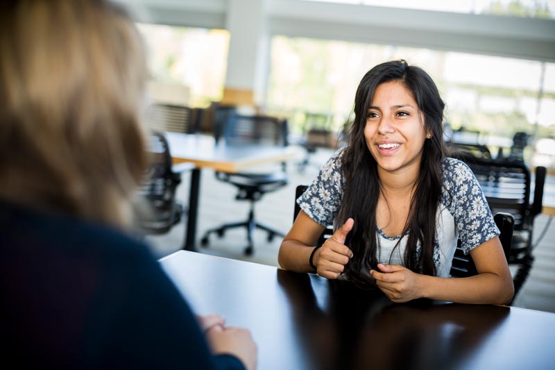 Two women sitting at a desk talking with each other.