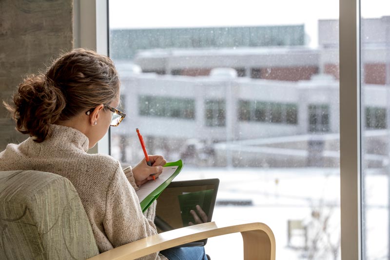 A girl sitting looking out of a large window while it is snowing outside.