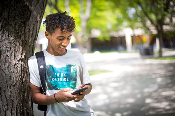 UVU student leaning against a tree on campus looking at phone
