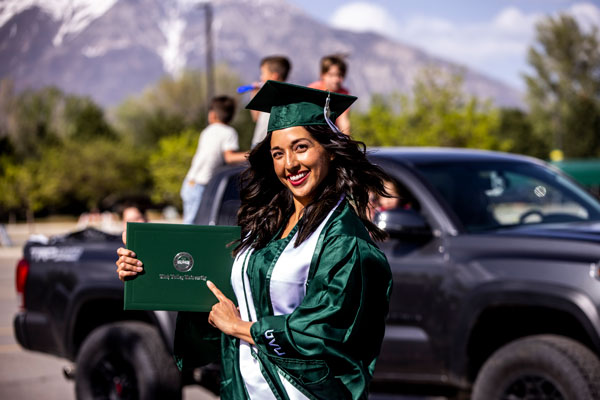 graduating UVU student dressed in cap and gown pointing at diploma and smiling