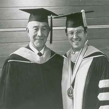 Wilson Sorensen standing with J. Marvin Higbee, both are wearing graduation regalia.