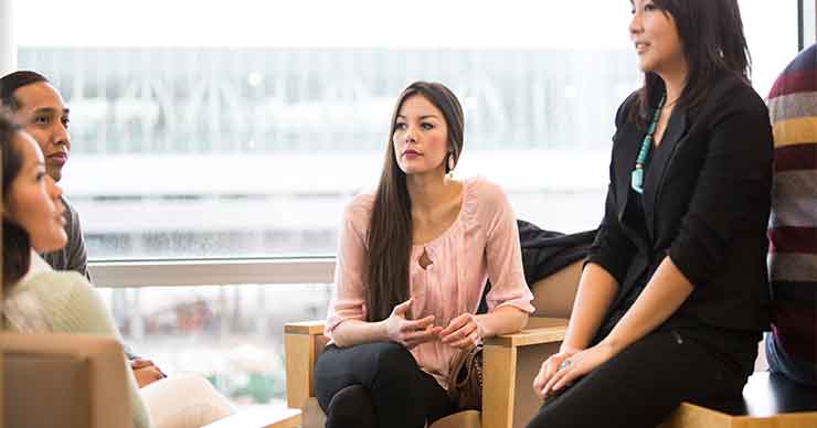 Group of students, focused on one female student sitting in a chair looking off in the distance.