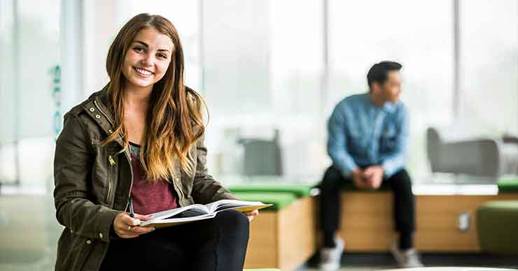 A female student sitting on a chair looking at a book with a male student reading in the background.