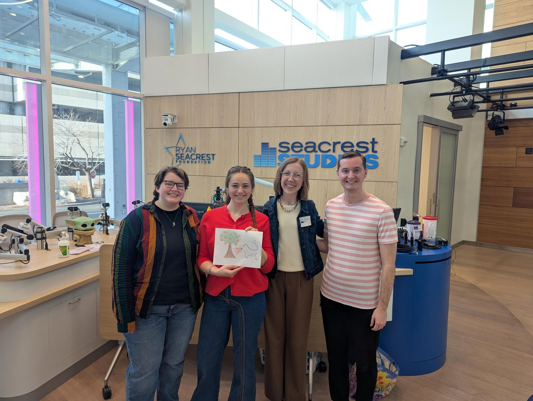 Four adults stand together in front of a broadcasting table, with the "Seacrest Studio" sign in the background. 
