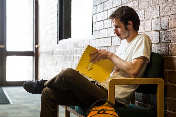 A man sitting on a chair looking a notebook he is holding.