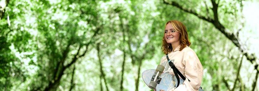 A woman walking through some trees with a skateboard in her arm.