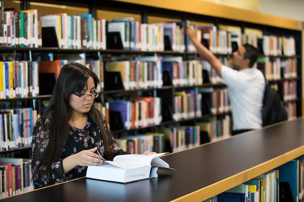 A woman reading a book and a man in the background looking through some book shelves.