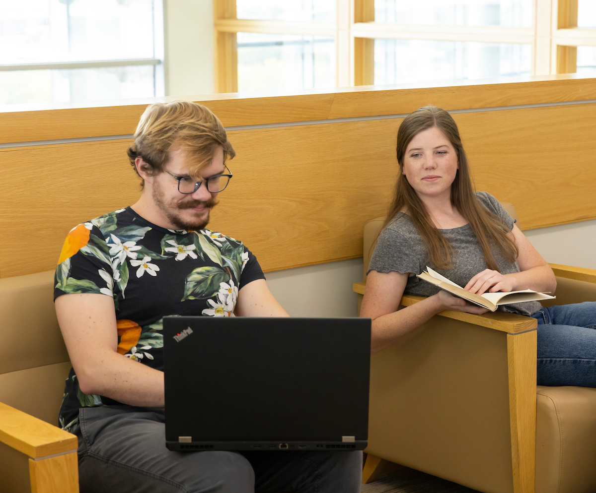 Image of a student on their laptop sitting next to a student reading a book