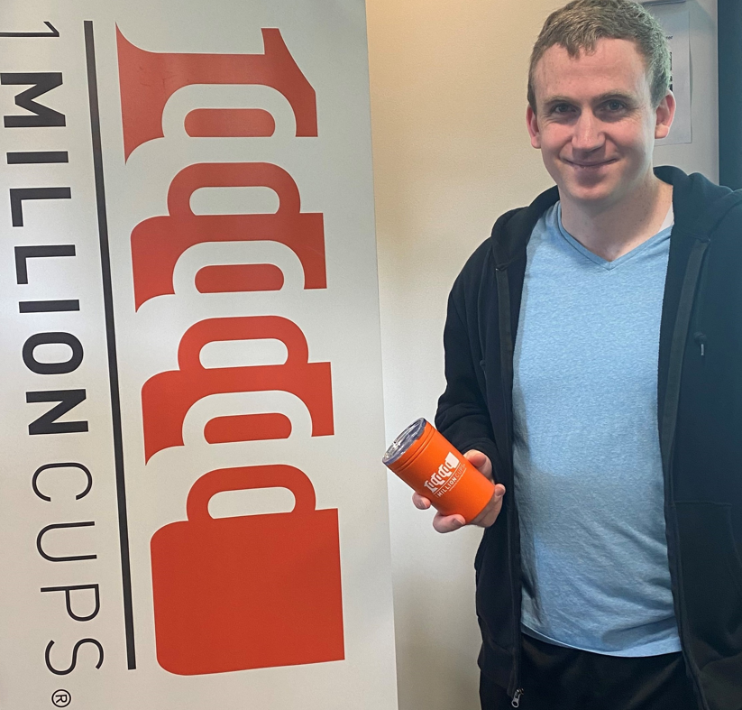 white man with light brown hair smiling and holding an orange 1 million cups insulated coffe cup next to the 1 Million Cups banner