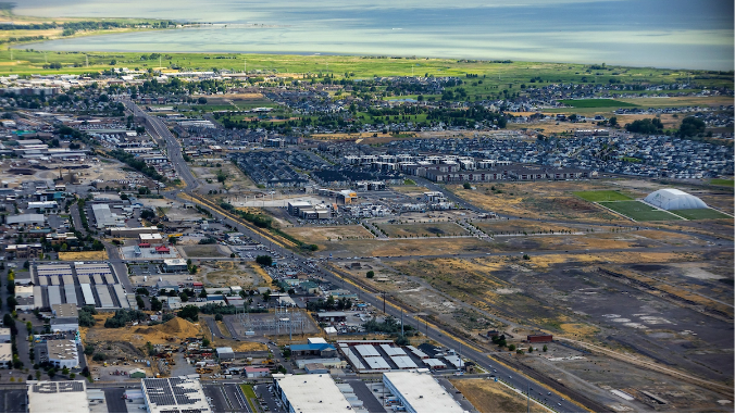 Aerial view of an urban landscape with industrial buildings, residential areas, greenery, and a body of water in the background.