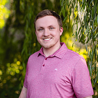 Alec Mathews wearing a pink shirt smiling outside