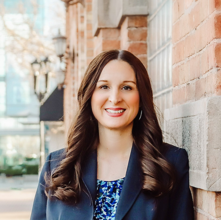 Dr. Kelly Hall is standing outside on a sunny day, smiling, facing forward, standing next to a brick wall.