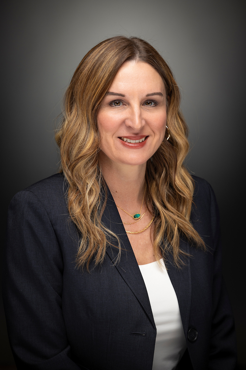 Dr. Kelly Hall is standing outside on a sunny day, smiling, facing forward, standing next to a brick wall.